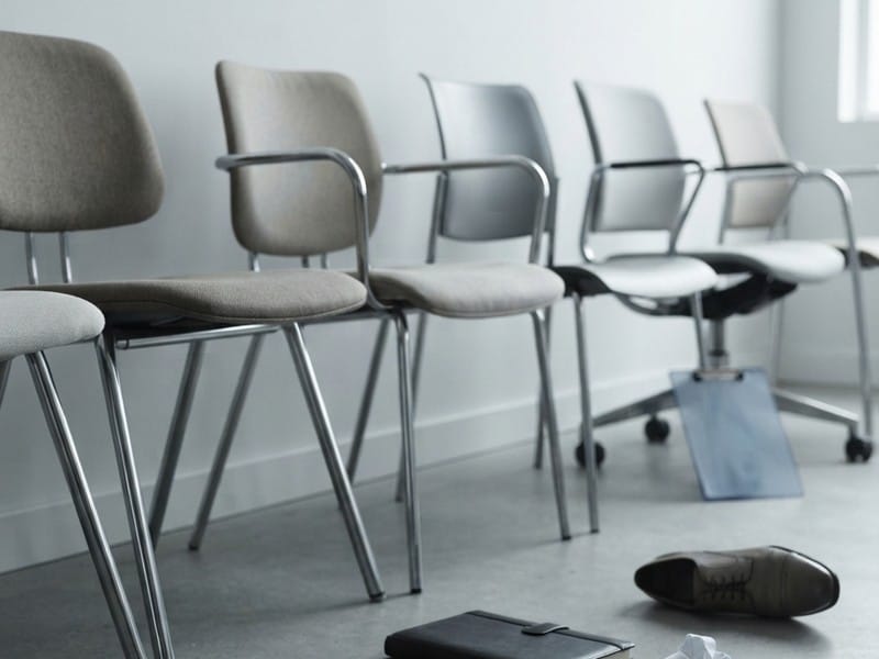 A row of empty office chairs lined up against a white wall, with a single dress shoe, a notebook, a clipboard, and crumpled paper lying on the floor.