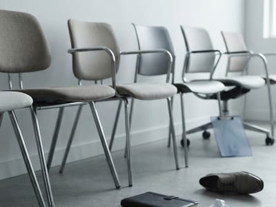A row of empty office chairs lined up against a white wall, with a single dress shoe, a notebook, a clipboard, and crumpled paper lying on the floor.
