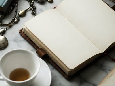 An open blank notebook in a leather cover rests on a marble table next to a cup of tea, a saucer, and beaded jewelry.